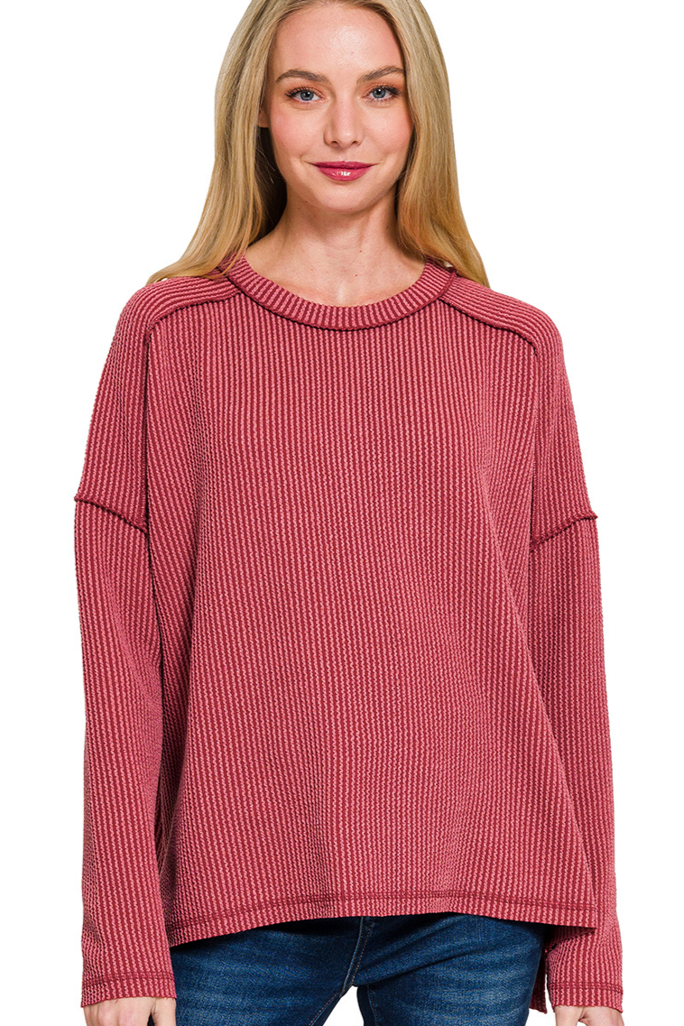 A burgundy ribbed boat neck top. The top has long sleeves, a relaxed fit, and raw edge details on the seams.