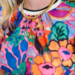 Vibrant floral print top in pink, orange, purple, and green with puff sleeves, round neckline, and contrast trim.