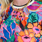 Vibrant floral print top in pink, orange, purple, and green with puff sleeves, round neckline, and contrast trim.