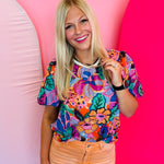 Vibrant floral print top in pink, orange, purple, and green with puff sleeves, round neckline, and contrast trim.