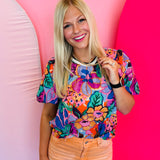 Vibrant floral print top in pink, orange, purple, and green with puff sleeves, round neckline, and contrast trim.