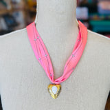 Close-up of a pink printed scarf-style necklace featuring a gold heart charm with a smooth white stone center, styled on a white background for a soft and elegant presentation.