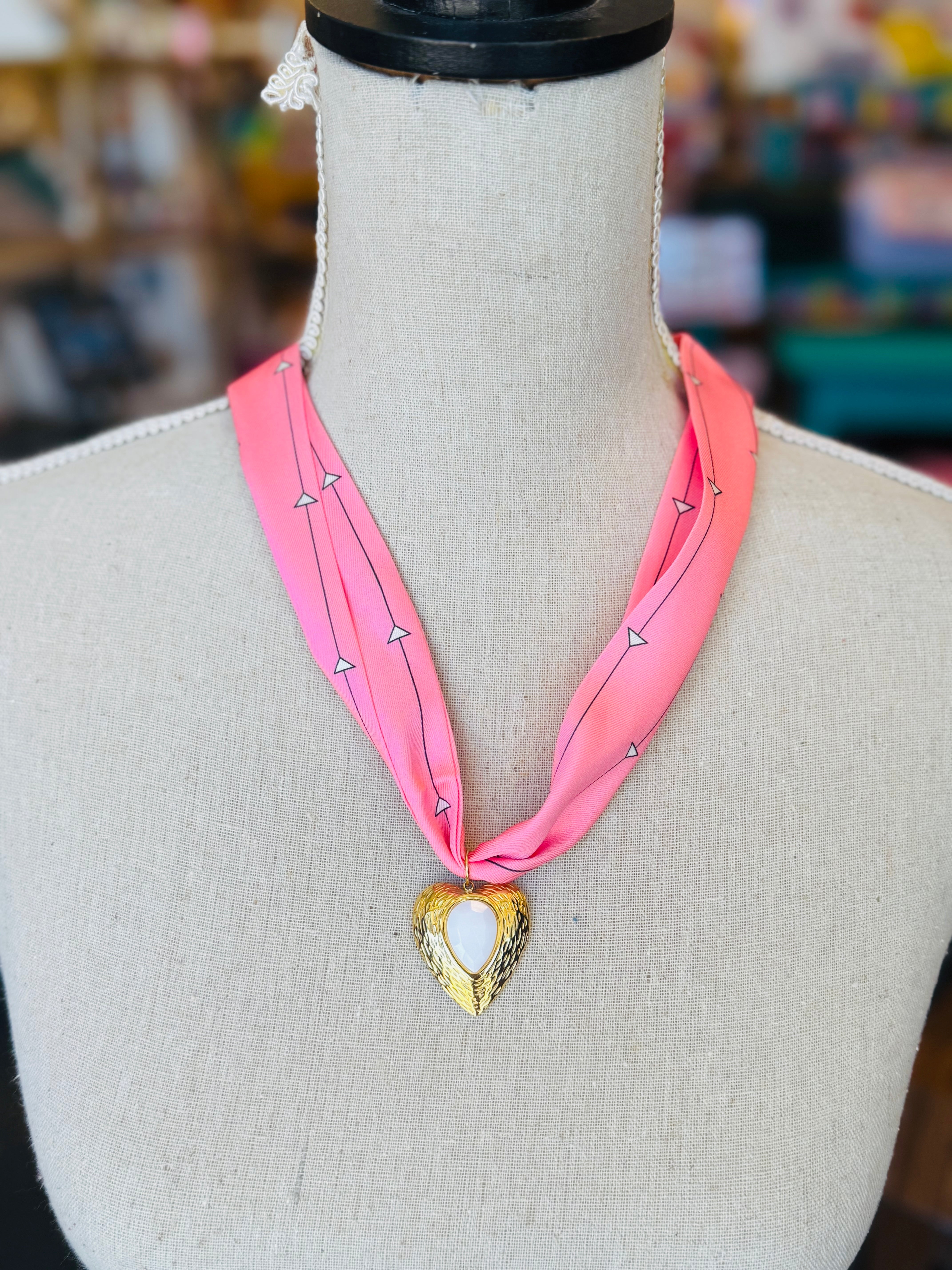 Close-up of a pink printed scarf-style necklace featuring a gold heart charm with a smooth white stone center, styled on a white background for a soft and elegant presentation.