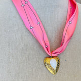 Close-up of a pink printed scarf-style necklace featuring a gold heart charm with a smooth white stone center, styled on a white background for a soft and elegant presentation.
