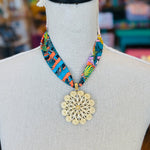 Tropical-inspired scarf necklace featuring a vibrant, multicolor fabric with jungle foliage patterns, accented by a shiny gold medallion charm at the center. Perfect for adding a bright, exotic flair to any outfit.