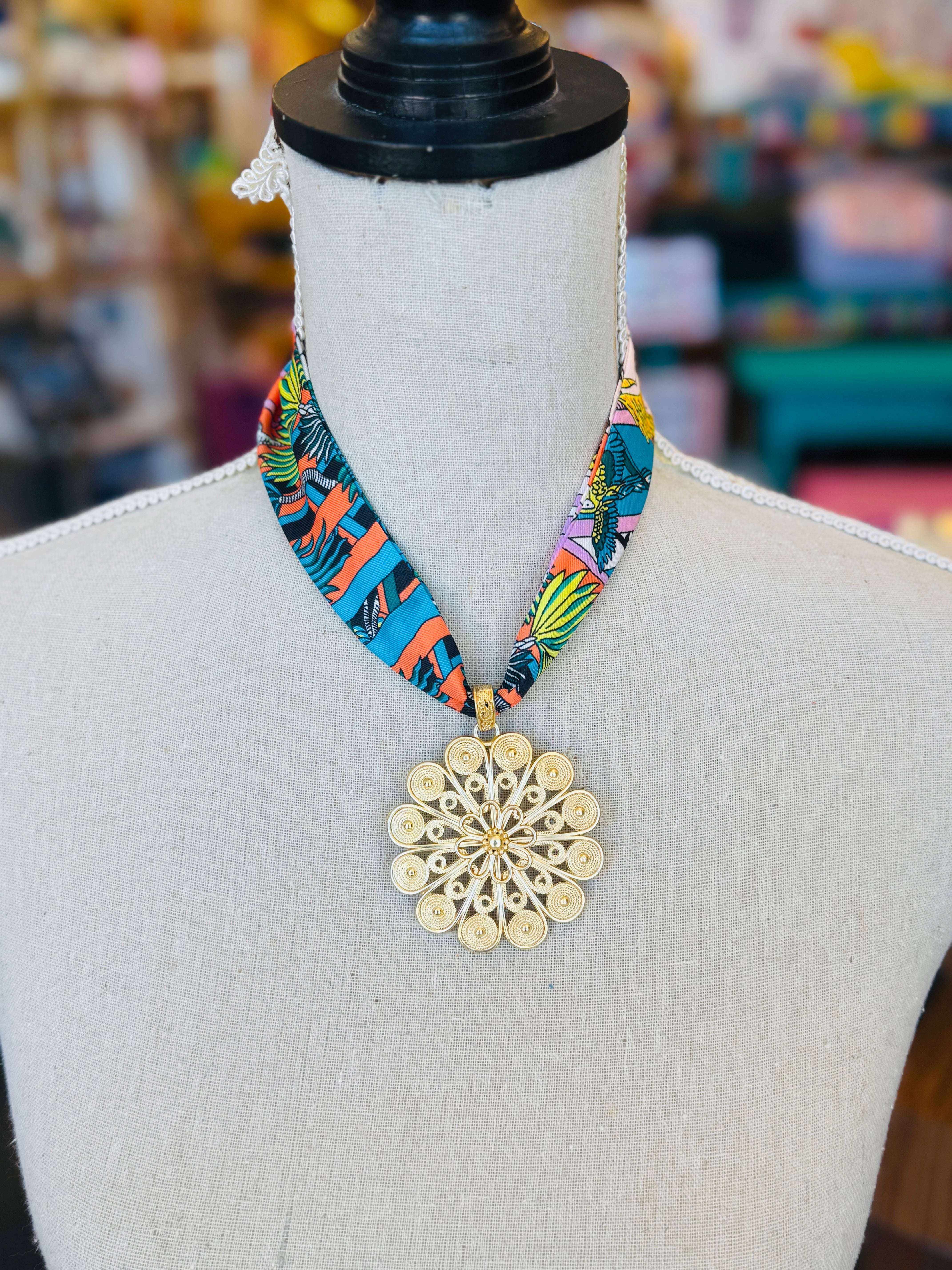 Tropical-inspired scarf necklace featuring a vibrant, multicolor fabric with jungle foliage patterns, accented by a shiny gold medallion charm at the center. Perfect for adding a bright, exotic flair to any outfit.