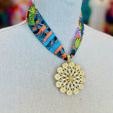 Tropical-inspired scarf necklace featuring a vibrant, multicolor fabric with jungle foliage patterns, accented by a shiny gold medallion charm at the center. Perfect for adding a bright, exotic flair to any outfit.