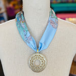 Blue printed scarf necklace with the words “lovely tiger,” featuring a gold cutout medallion charm; a playful and whimsical accessory perfect for adding a touch of charm and elegance to any outfit.
