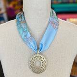 Blue printed scarf necklace with the words “lovely tiger,” featuring a gold cutout medallion charm; a playful and whimsical accessory perfect for adding a touch of charm and elegance to any outfit.
