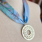Blue printed scarf necklace with the words “lovely tiger,” featuring a gold cutout medallion charm; a playful and whimsical accessory perfect for adding a touch of charm and elegance to any outfit.