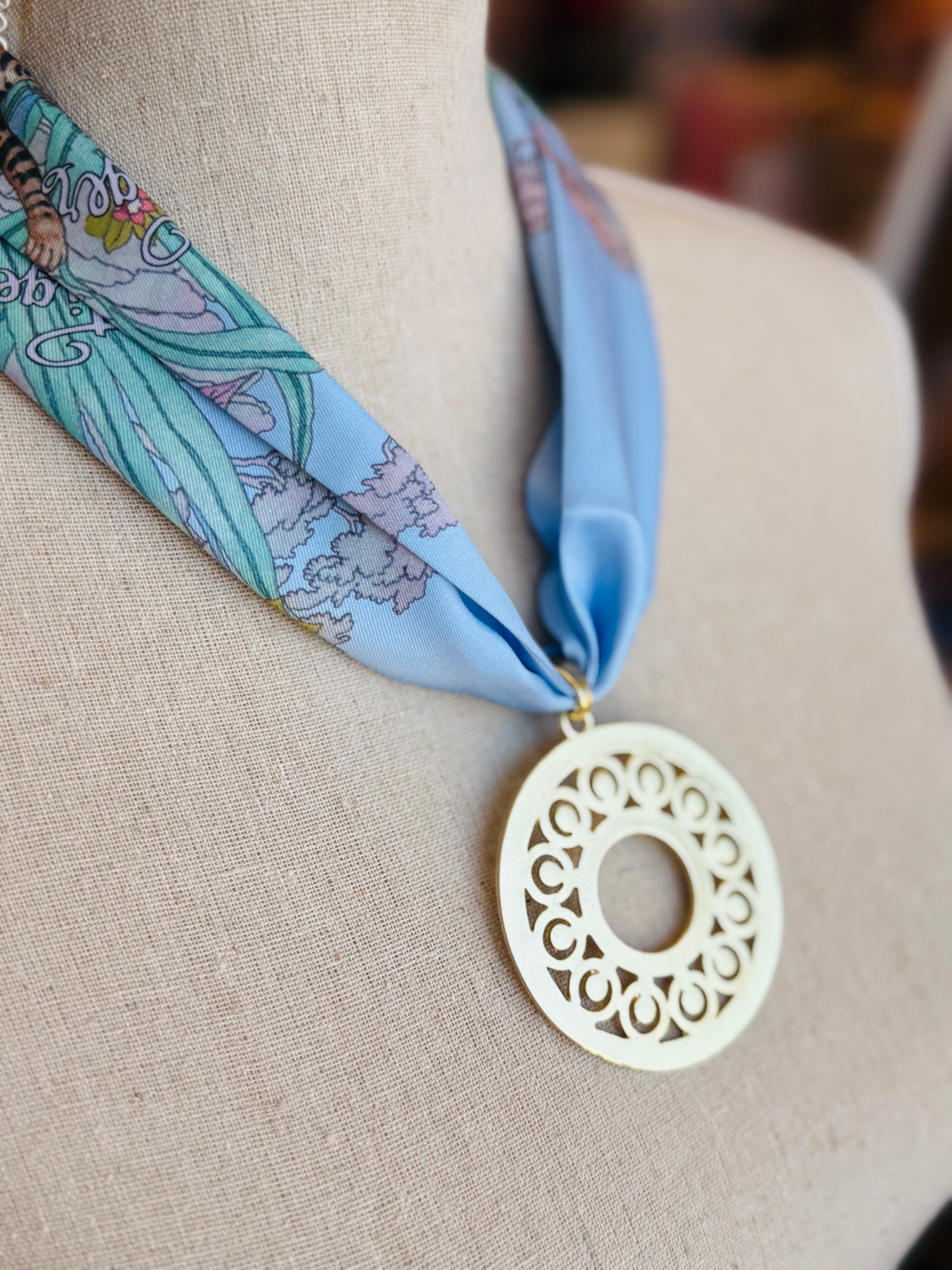 Blue printed scarf necklace with the words “lovely tiger,” featuring a gold cutout medallion charm; a playful and whimsical accessory perfect for adding a touch of charm and elegance to any outfit.