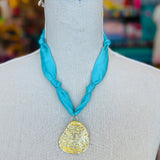 Close-up image of a blue scarf-style necklace with a textured gold medallion charm, styled to evoke breezy coastal vibes. The soft aqua blue fabric contrasts elegantly with the bold gold pendant, creating a light and stylish accessory perfect for casual or chic outfits.