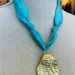 Close-up image of a blue scarf-style necklace with a textured gold medallion charm, styled to evoke breezy coastal vibes. The soft aqua blue fabric contrasts elegantly with the bold gold pendant, creating a light and stylish accessory perfect for casual or chic outfits.