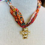 Scarf necklace featuring a richly patterned fabric band and a sculpted gold cross charm with sparkling crystal accents, styled to add vintage charm and elegance to any outfit.
