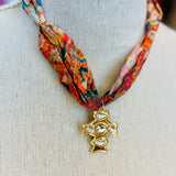 Scarf necklace featuring a richly patterned fabric band and a sculpted gold cross charm with sparkling crystal accents, styled to add vintage charm and elegance to any outfit.