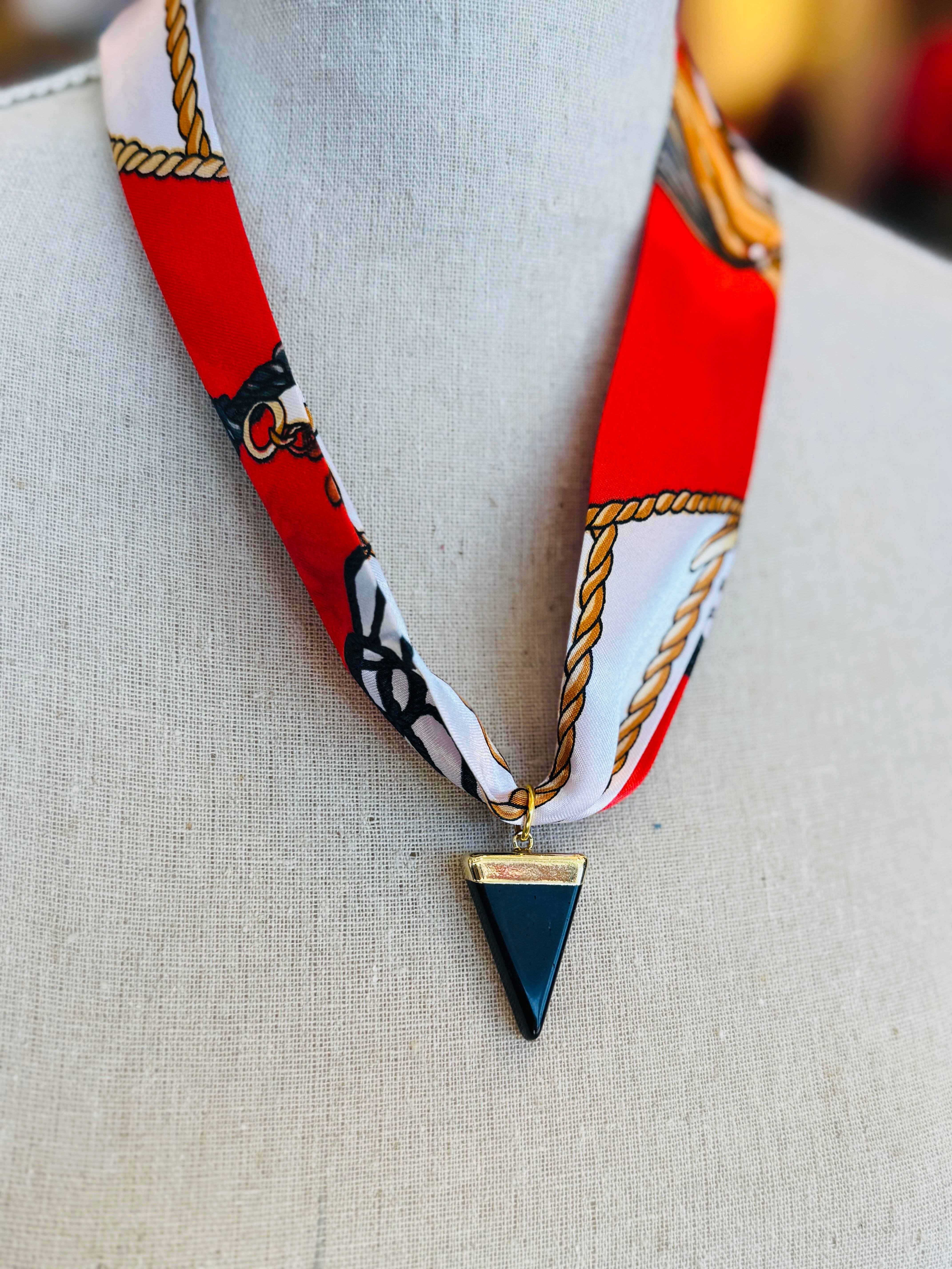 Red patterned scarf necklace featuring a sleek black triangle pendant with gold trim, styled to add a bold yet elegant statement to any outfit.