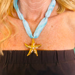 Blue Print Scarf Starfish Charm Necklace featuring a soft aqua scarf band paired with a sparkling gold starfish pendant, accented with delicate pearls and shimmer. A playful, beach-inspired accessory perfect for adding a coastal touch to any outfit.