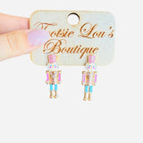 Pastel nutcracker earrings with pink hats, teal pants, gold sparkle trim, and cheerful faces.