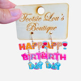 A pair of playful dangle earrings featuring colorful charms, including letter beads that spell out "Happy Birthday," a miniature party hat, and a small wine glass.