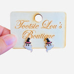 A pair of stud earrings shaped like small, white cartoon ghosts. Each ghost is wearing a glittery black witch hat that has a small pink accent band.