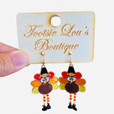 A pair of dangle earrings shaped like turkeys. The turkeys have colorful enamel tail feathers, are wearing small black pilgrim hats, and have little legs that dangle.