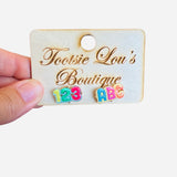 A pair of stud earrings with a white base. One earring features the letters "ABC" in colorful enamel, and the other features the numbers "123" in colorful enamel.