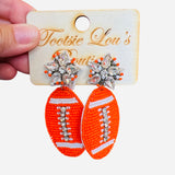A pair of dangle earrings, each with a sparkling crystal star stud at the top. Dangling below is a football-shaped charm made from vibrant orange beads.