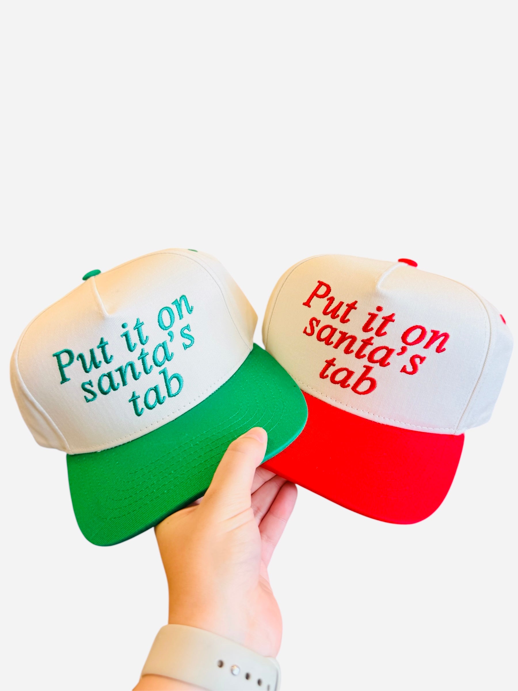 Red and green baseball hats with “Put It On Santa’s Tab” embroidery, festive Christmas accessory.