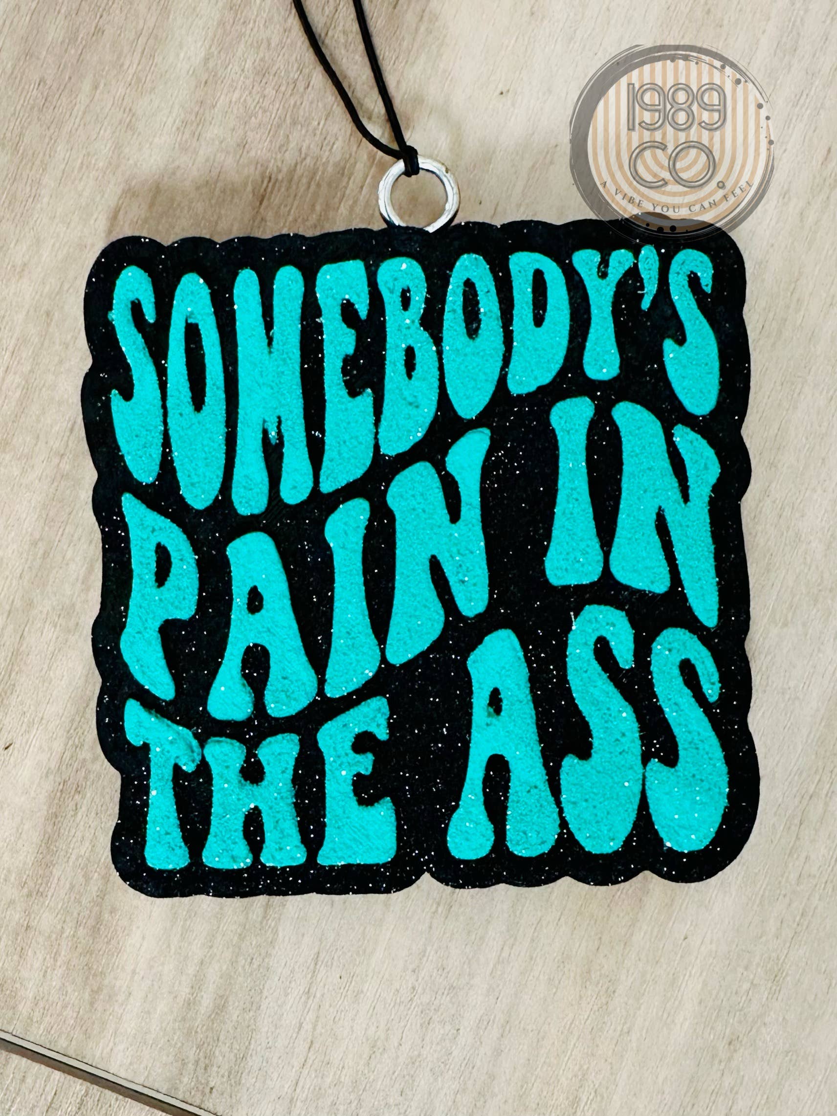 Black car freshie with teal text reading “Somebody’s Pain In The Ass.”