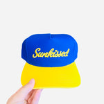 Blue hat with yellow bill and “Sunkissed” embroidered on front, adjustable strap in back