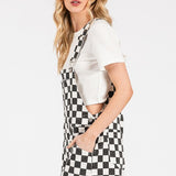 Black checkered knit romper overalls with cuffed shorts, chest and side pockets, and adjustable straps, laid flat to show texture and structure.