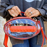 Clear football-shaped crossbody bag with orange trim, gold hardware, top handle, and adjustable strap, displayed to show shape and transparency.