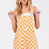 Orange checkered knit romper overalls with cuffed shorts, adjustable straps, and multiple pockets, laid flat to show texture and design.
