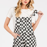 Black checkered knit romper overalls with cuffed shorts, chest and side pockets, and adjustable straps, laid flat to show texture and structure.