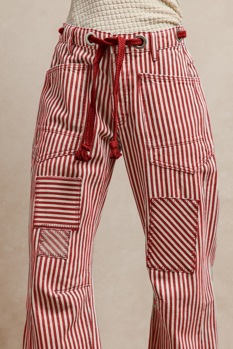 Red striped twill barrel-leg pants with bold vertical stripes, featuring a red rope tie threaded through silver eyelets and decorative front pockets; made from 100% cotton for breathable comfort and standout style.