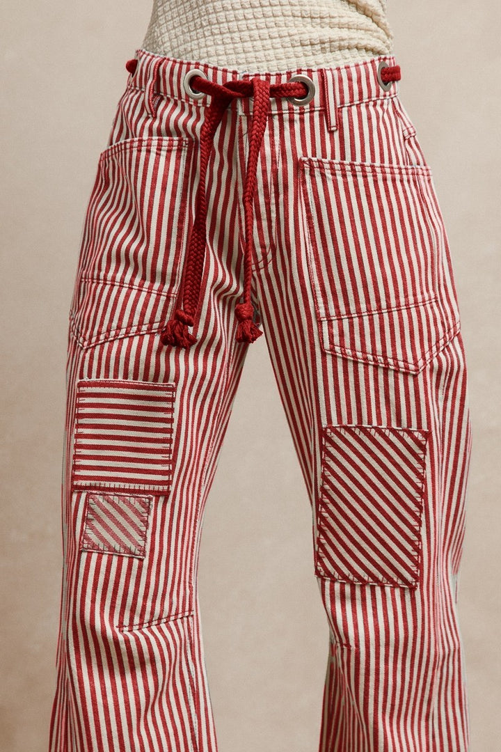 Red striped twill barrel-leg pants with bold vertical stripes, featuring a red rope tie threaded through silver eyelets and decorative front pockets; made from 100% cotton for breathable comfort and standout style.