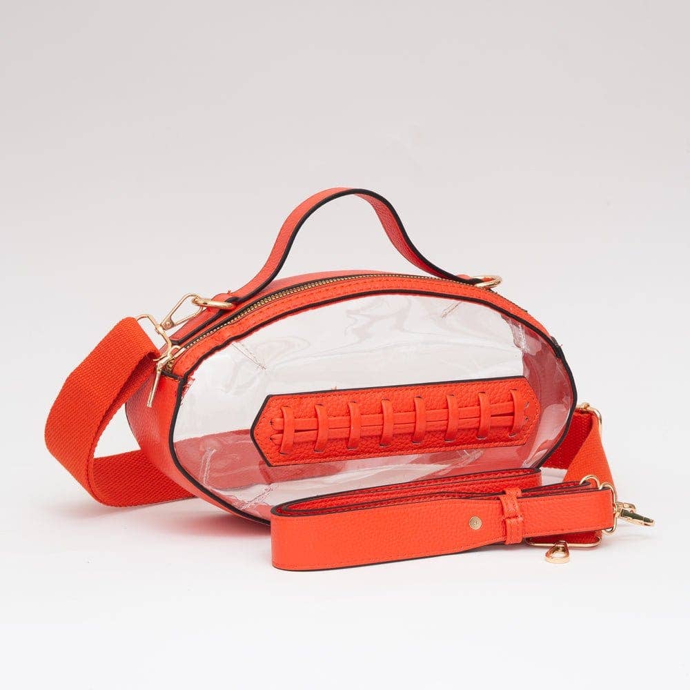 Clear football-shaped crossbody bag with orange trim, gold hardware, top handle, and adjustable strap, displayed to show shape and transparency.