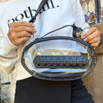 Clear football-shaped crossbody bag with black trim, gold hardware, top handle, and adjustable strap, laid flat to highlight shape and detailing.