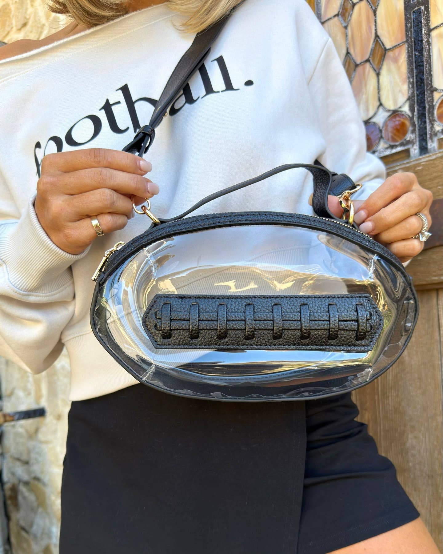 Clear football-shaped crossbody bag with black trim, gold hardware, top handle, and adjustable strap, laid flat to highlight shape and detailing.