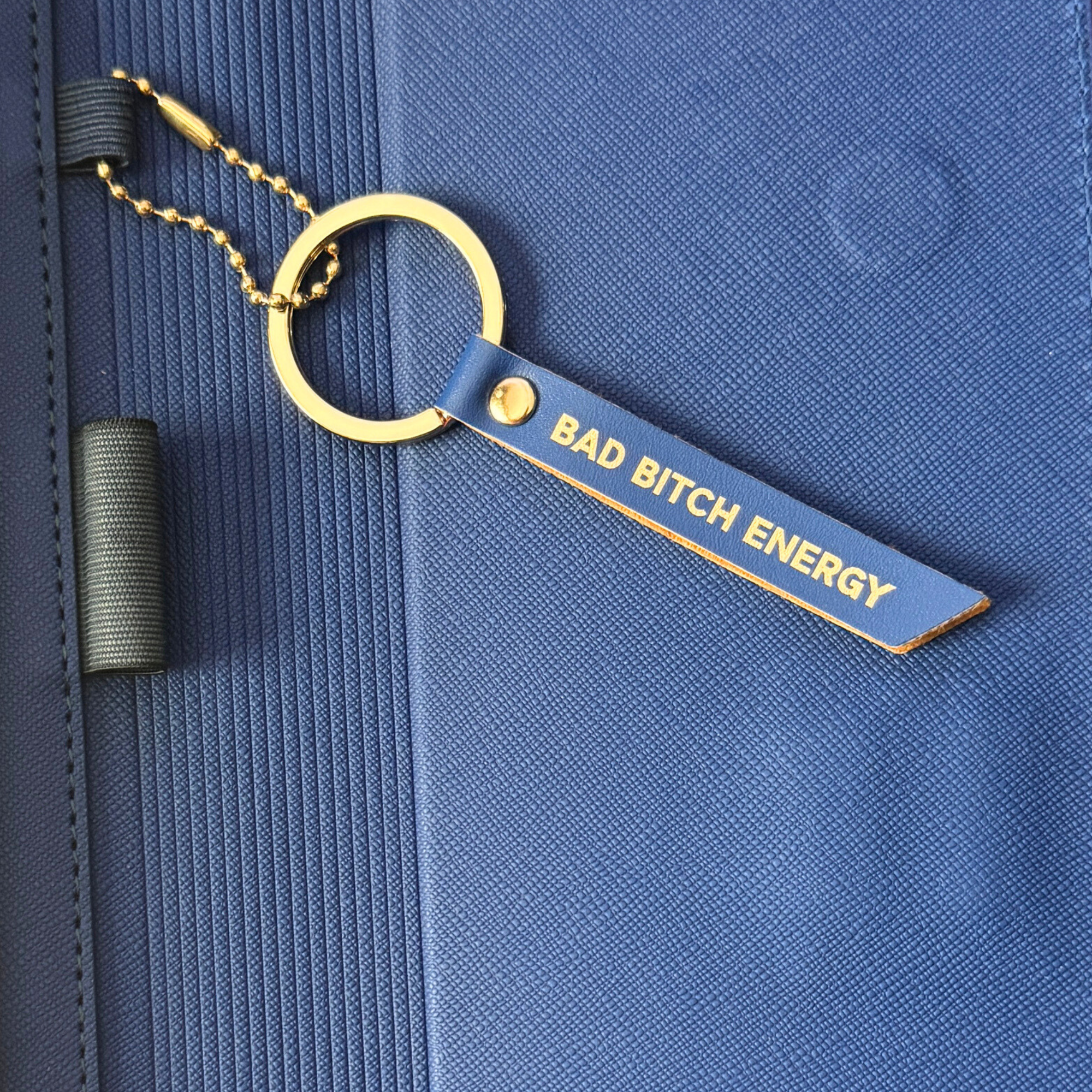 Keychain with 'Bad Bitch Energy' tag attached to a blue folio.