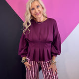 Burgundy peplum top with pleated waist, round neckline, and long balloon sleeves.
