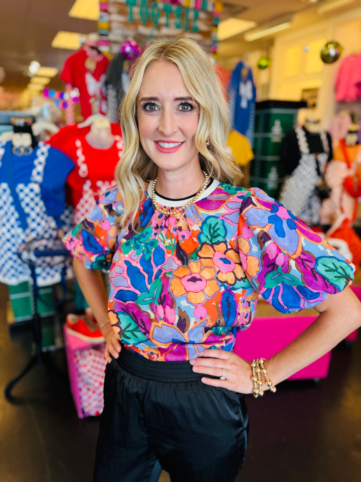 Vibrant floral print top in pink, orange, purple, and green with puff sleeves, round neckline, and contrast trim.