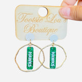 Gold hoops with green glitter dangles and white writing that says Hawks set on a white stud.