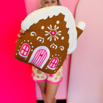 Gingerbread house pillow with frosting details and pink candy accents, soft festive holiday decor.