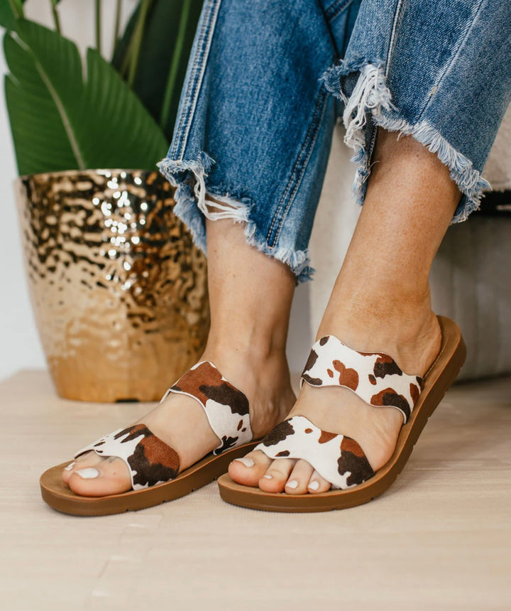 Brown cow print Corkys Wishy Washy sandals featuring playful crisscross straps, cushioned footbed, and a comfortable, modern slip-on design perfect for casual summer wear.