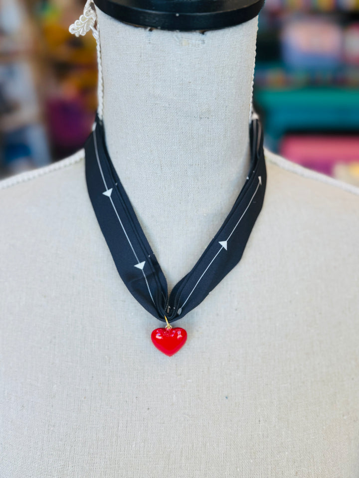 A black scarf necklace with delicate white patterns, accented by a bold red heart charm for a striking contrast. A lightweight, stylish piece perfect for adding a pop of personality to any outfit.