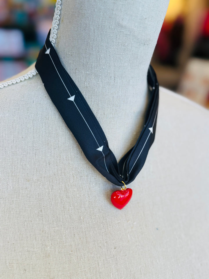 A black scarf necklace with delicate white patterns, accented by a bold red heart charm for a striking contrast. A lightweight, stylish piece perfect for adding a pop of personality to any outfit.
