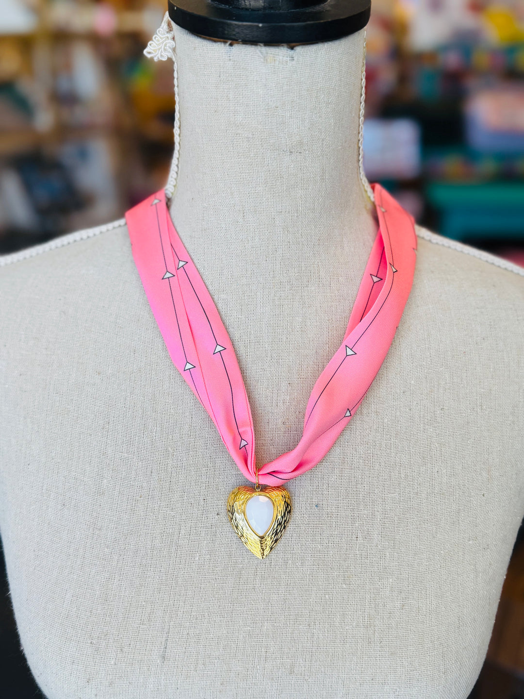 Close-up of a pink printed scarf-style necklace featuring a gold heart charm with a smooth white stone center, styled on a white background for a soft and elegant presentation.