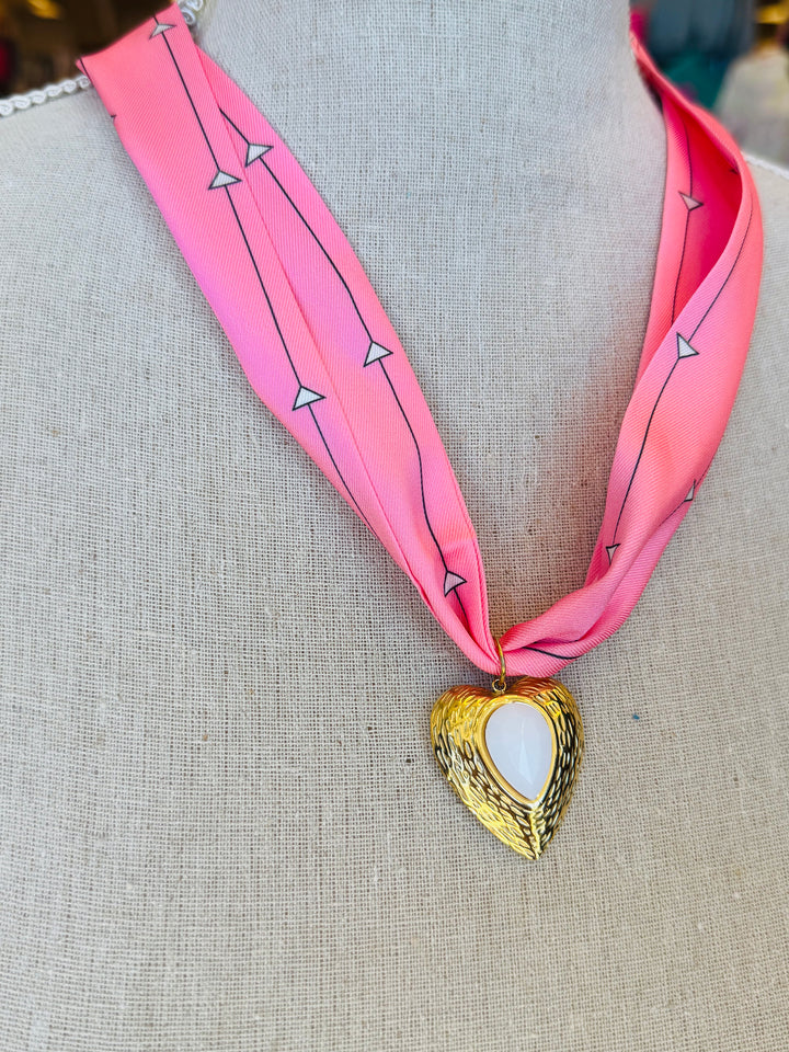 Close-up of a pink printed scarf-style necklace featuring a gold heart charm with a smooth white stone center, styled on a white background for a soft and elegant presentation.