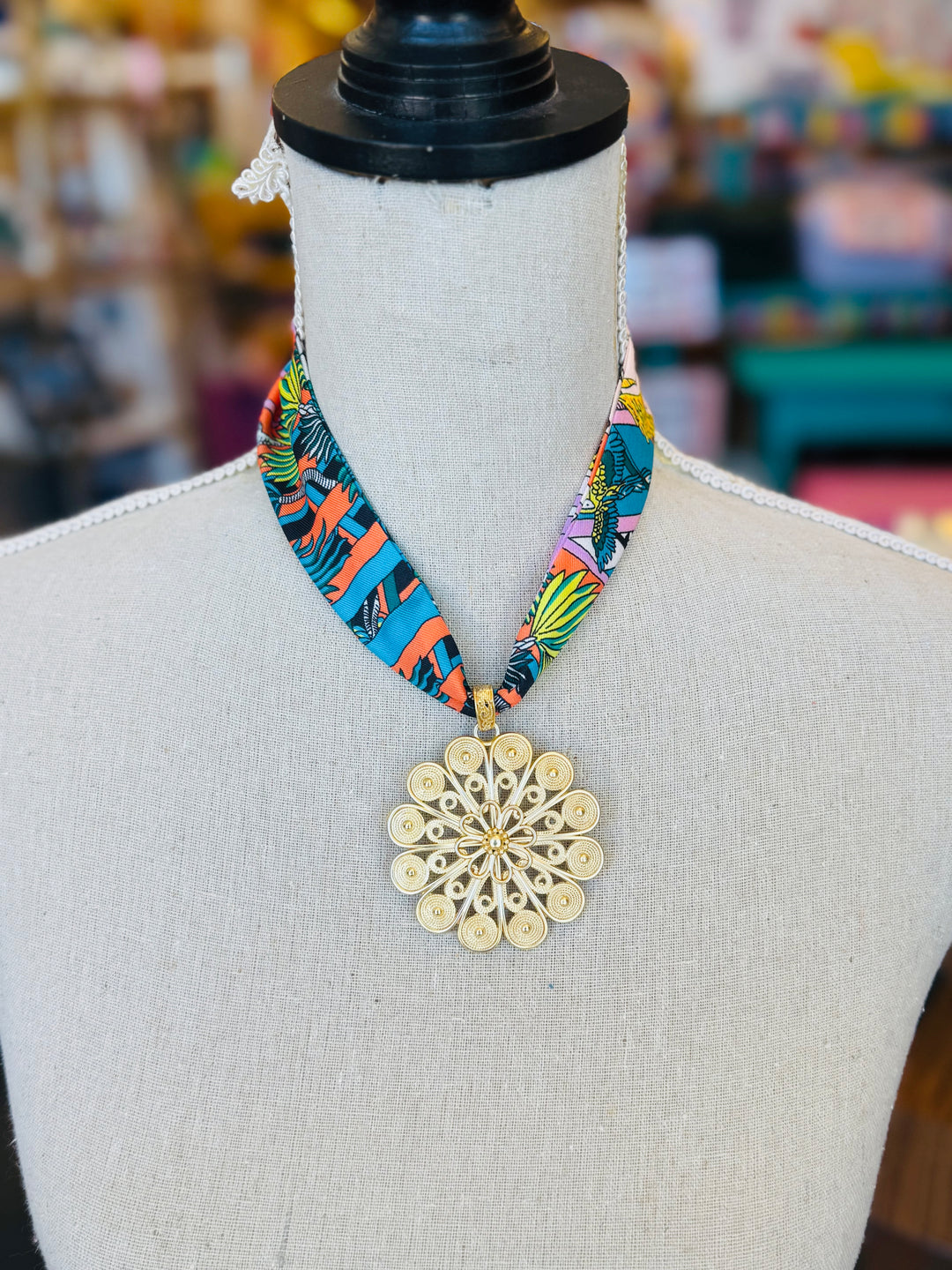 Tropical-inspired scarf necklace featuring a vibrant, multicolor fabric with jungle foliage patterns, accented by a shiny gold medallion charm at the center. Perfect for adding a bright, exotic flair to any outfit.